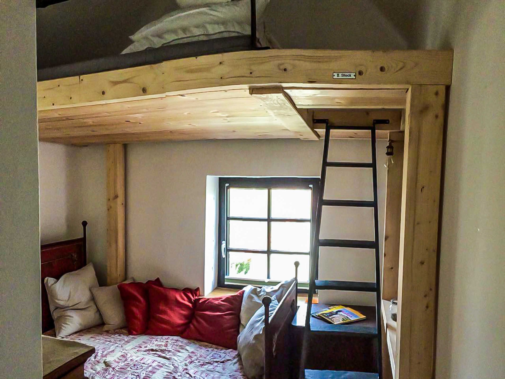 A compact room with a bunk bed, a desk with a chair, a window, and a ladder.