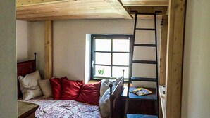 Ein kleines Zimmer mit Etagenbett, Schreibtisch und Stuhl, einem Fenster und einer Leitern.