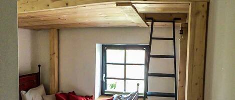 A compact room with a bunk bed, a desk with a chair, a window, and a ladder.