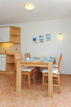 Ein Essbereich mit Holztisch und -stühlen, eine Küche mit weißen Schränken und drei kleine Bilder mit Strandmotiv an der Wand.