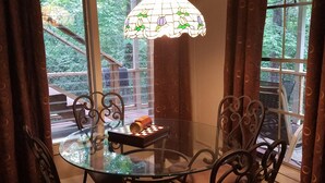 A dining area with a glass table and metal chairs, a hanging light fixture, and a view of the outdoors.