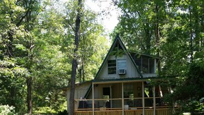 A cabin with a wooden deck and a covered porch, surrounded by dense forest.