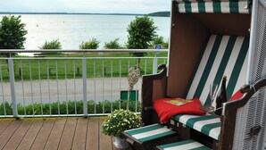 Ein Korbsessel mit gestreifter Sitzauflage und einem roten Kissen, ein kleiner Tisch mit Glas und Flasche, und ein Blumentopf auf einer Holzterrasse mit Blick auf ein Gewässer.