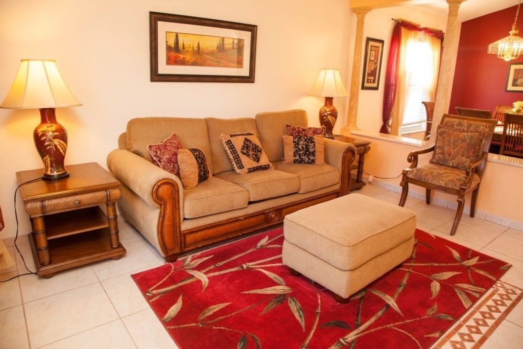 A living room with a sofa, ottoman, and side tables, featuring a red patterned rug and framed artwork on the walls.