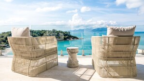 Deux fauteuils en osier avec des coussins, une petite table en pierre et une balustrade en verre donnant sur une mer turquoise et un paysage verdoyant.