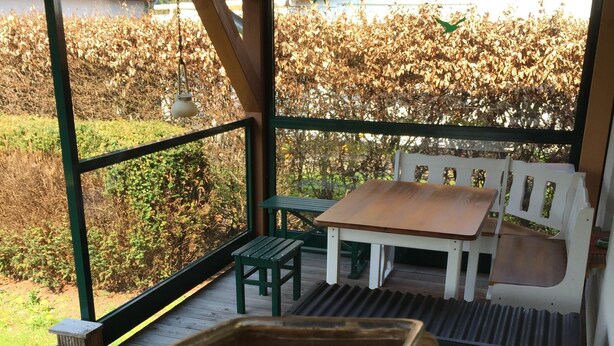 Un espace extérieur abrité, avec une table et des bancs en bois, un petit tapis et une pancarte de bienvenue.