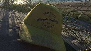 Une casquette jaune, ornée d’une inscription, est posée sur une surface sablonneuse, avec de l’herbe au premier plan et une plage en arrière-plan.