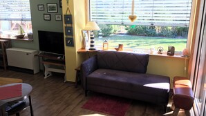 Un salon confortable avec un canapé, un téléviseur, une petite table et une fenêtre équipée de stores.