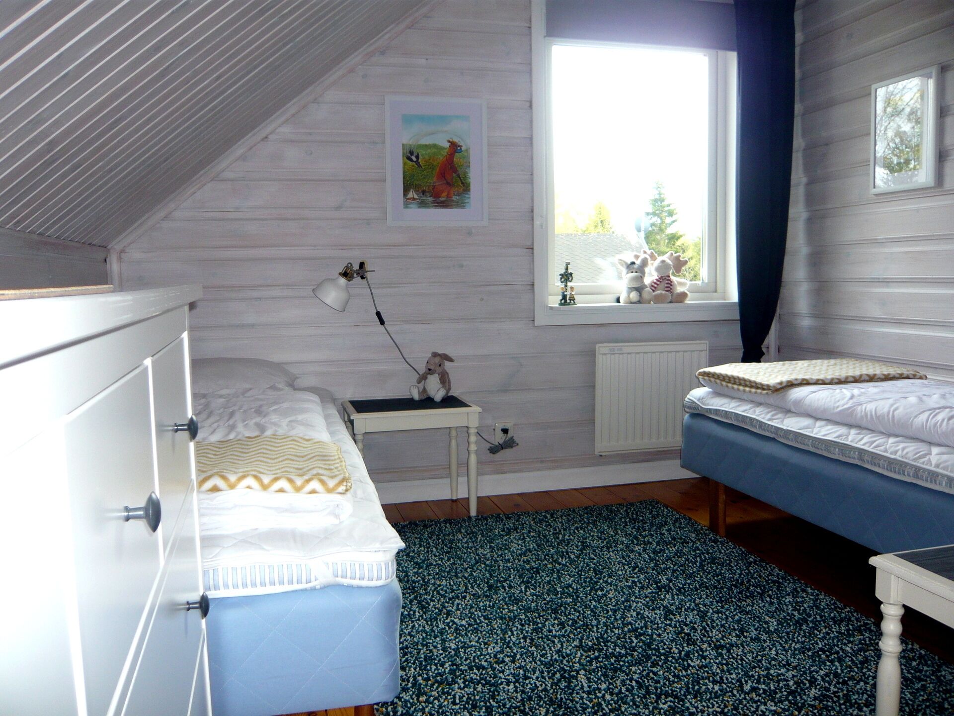 Ein Schlafzimmer mit zwei Betten, einem Fenster mit Blick auf Bäume, einem Heizkörper und einem kleinen Tisch mit einer Lampe und Stofftieren.