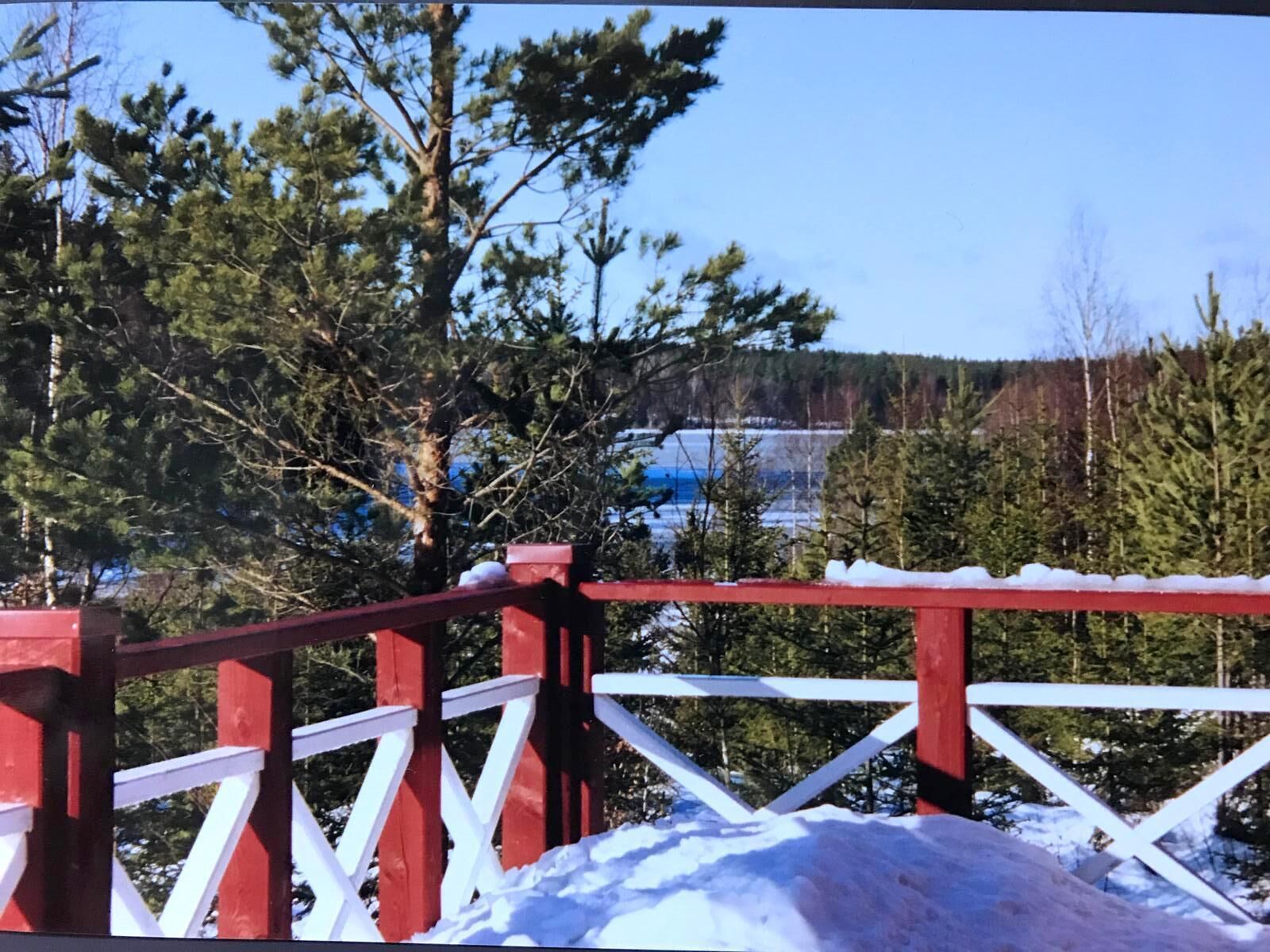 Una cerca de madera roja y blanca con nieve, que da vista a un bosque y una masa de agua.