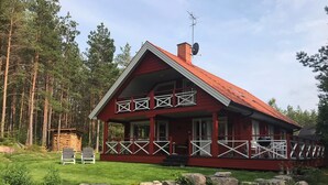 A red wooden cabin with a porch, surrounded by a stone wall and a forest.
