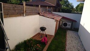 Une cour arrière avec une clôture en bois, un barbecue, un petit jardin fleuri et un mur blanc.