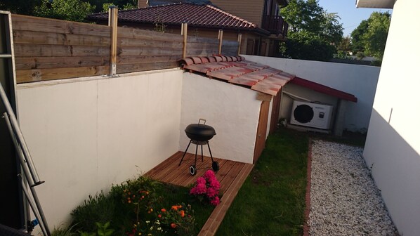 A backyard with a wooden fence, a barbecue grill, a small garden with flowers, and a white wall.