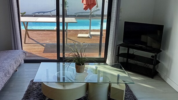 Un salon avec une table basse en verre, un canapé, un téléviseur et une vue sur une piscine et une plage, le tout visible à travers de grandes fenêtres.