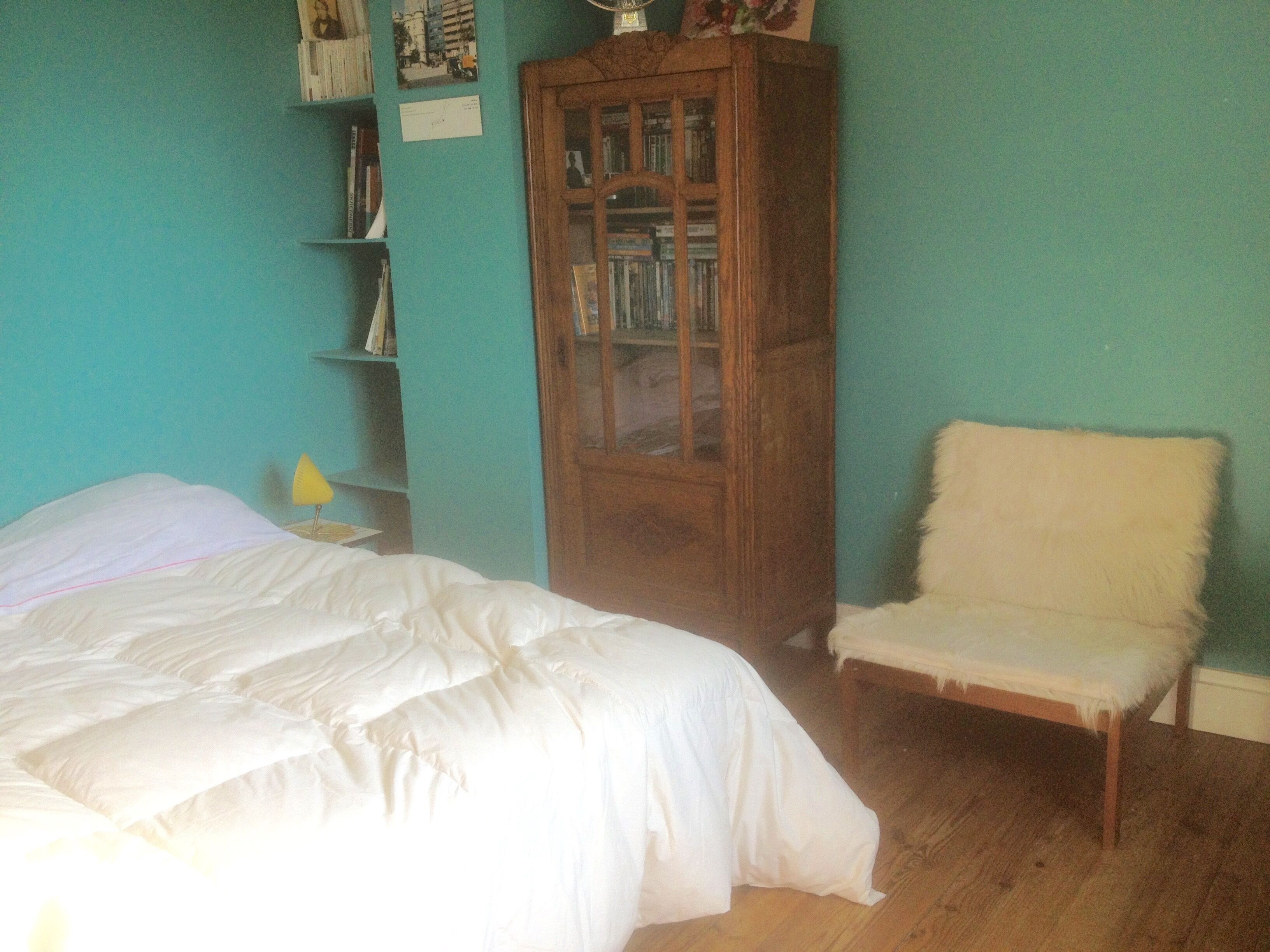 Ein Schlafzimmer mit einem Bett, einem Sessel, einer Holzkommode und einem Bücherregal vor einer türkisblauen Wand.