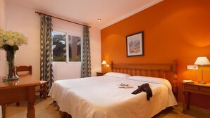 Une chambre d’hôtel avec un grand lit, une tête de lit en bois, deux lampes de chevet, une fenêtre avec des rideaux, un bureau agrémenté d’un vase de fleurs et un tableau encadré au mur.