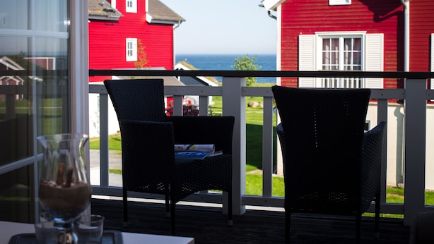 Un balcon avec des fauteuils en osier, une table et une vue sur des maisons rouges et l’océan.