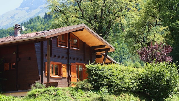 A wooden cabin with a sloped roof, large windows, and a chimney, surrounded by lush greenery and mountains in the background. - Typical Savoyard chalet, independent, warm and cozy (Thônes)