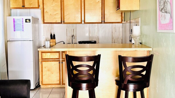 A kitchen with wooden cabinets, a white refrigerator, a sink, and two wooden chairs. - Queen apartment with generator, washing machine & patio furniture for relaxation (Nassau)