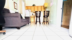 A modern living room with a grey sofa, wooden ceiling fan, and a kitchen area with wooden cabinets and two bar stools. - Queen apartment with generator, washing machine & patio furniture for relaxation (Nassau)