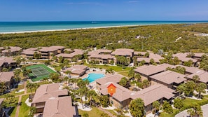 A resort with multiple bungalow-style buildings, a central swimming pool, tennis courts, and a beach view.