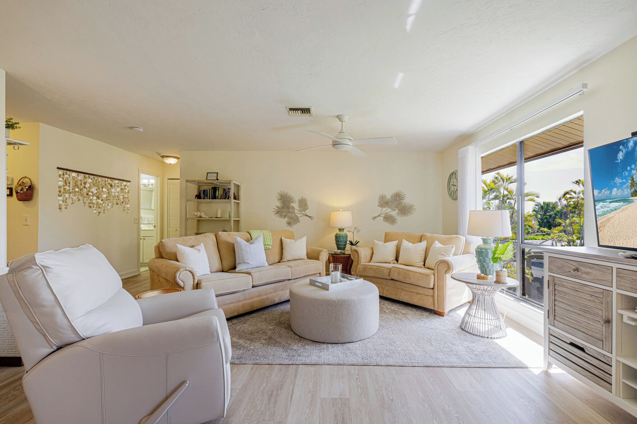 A living room with a beige sofa set, a round ottoman, a small side table, and a television on a wooden cabinet.