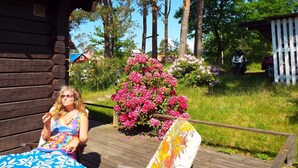 A wooden deck with a person sitting on a chair, surrounded by blooming pink flowers and a wooden cabin in the background. - Fantastic family vacation 600m from sandy beach, large garden, near forest, river (Mellbystrand)