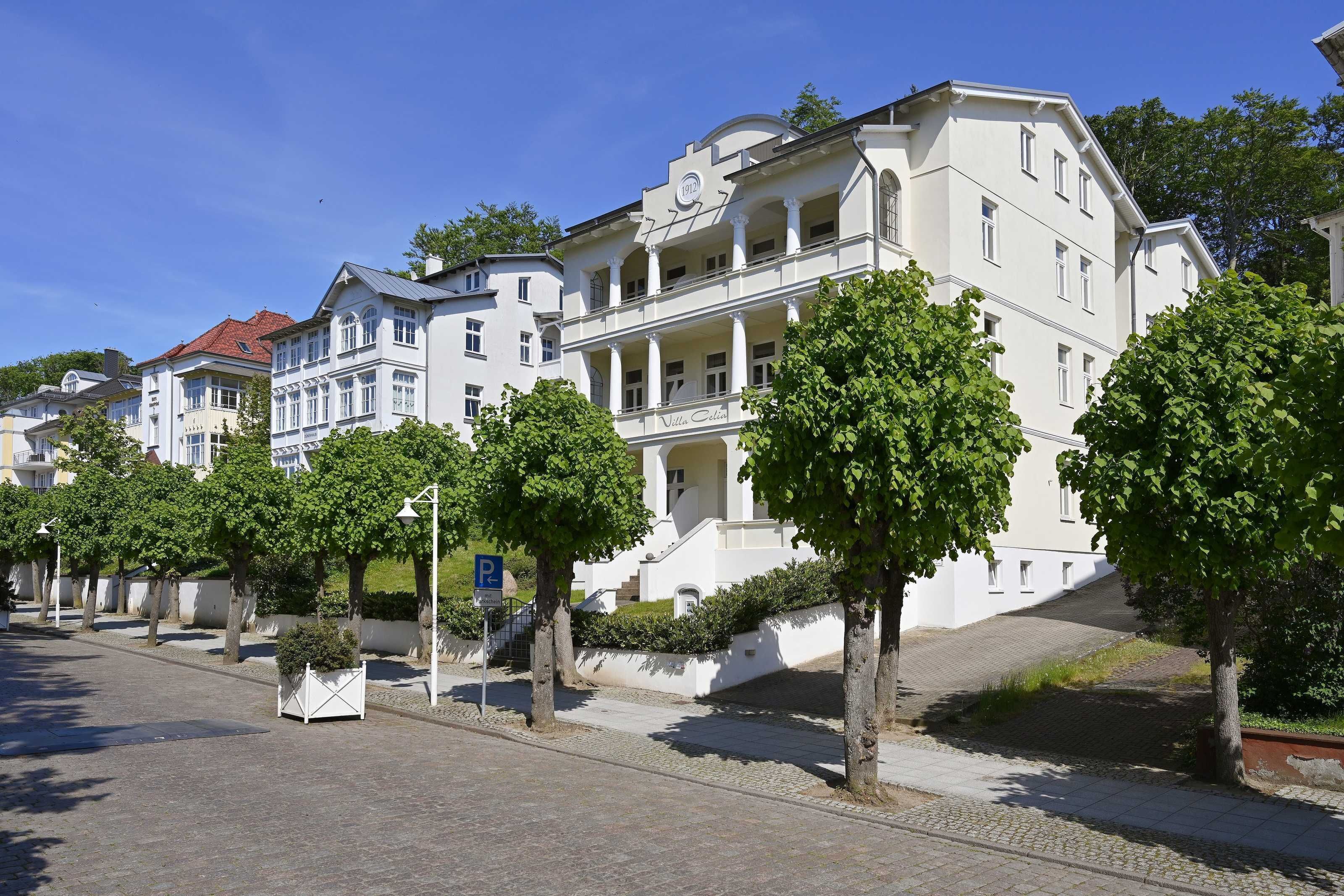 Reihenweise weiße Gebäude mit Balkonen, ein klarer, blauer Himmel und Bäume säumen die Straße.