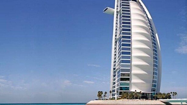 Um edifício alto e cilíndrico com uma estrutura em forma de vela, situado perto de uma praia com águas azul-claras.