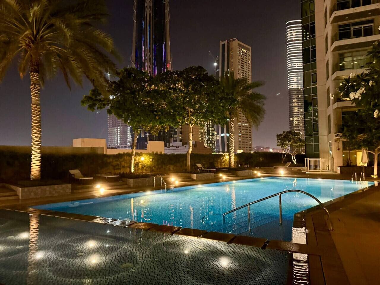 A swimming pool with a cityscape view at night, surrounded by palm trees and modern buildings.