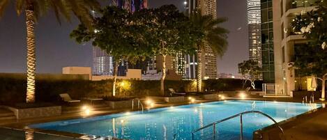 A swimming pool with a cityscape view at night, surrounded by palm trees and modern buildings.