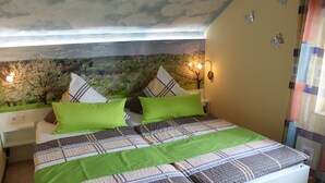 A bedroom with a bed, green pillows, a nightstand, a wall-mounted lamp, and a mural of a landscape.