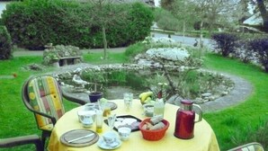 A round table with a yellow tablecloth, surrounded by chairs with striped cushions, set in a garden with a pond and stone features.