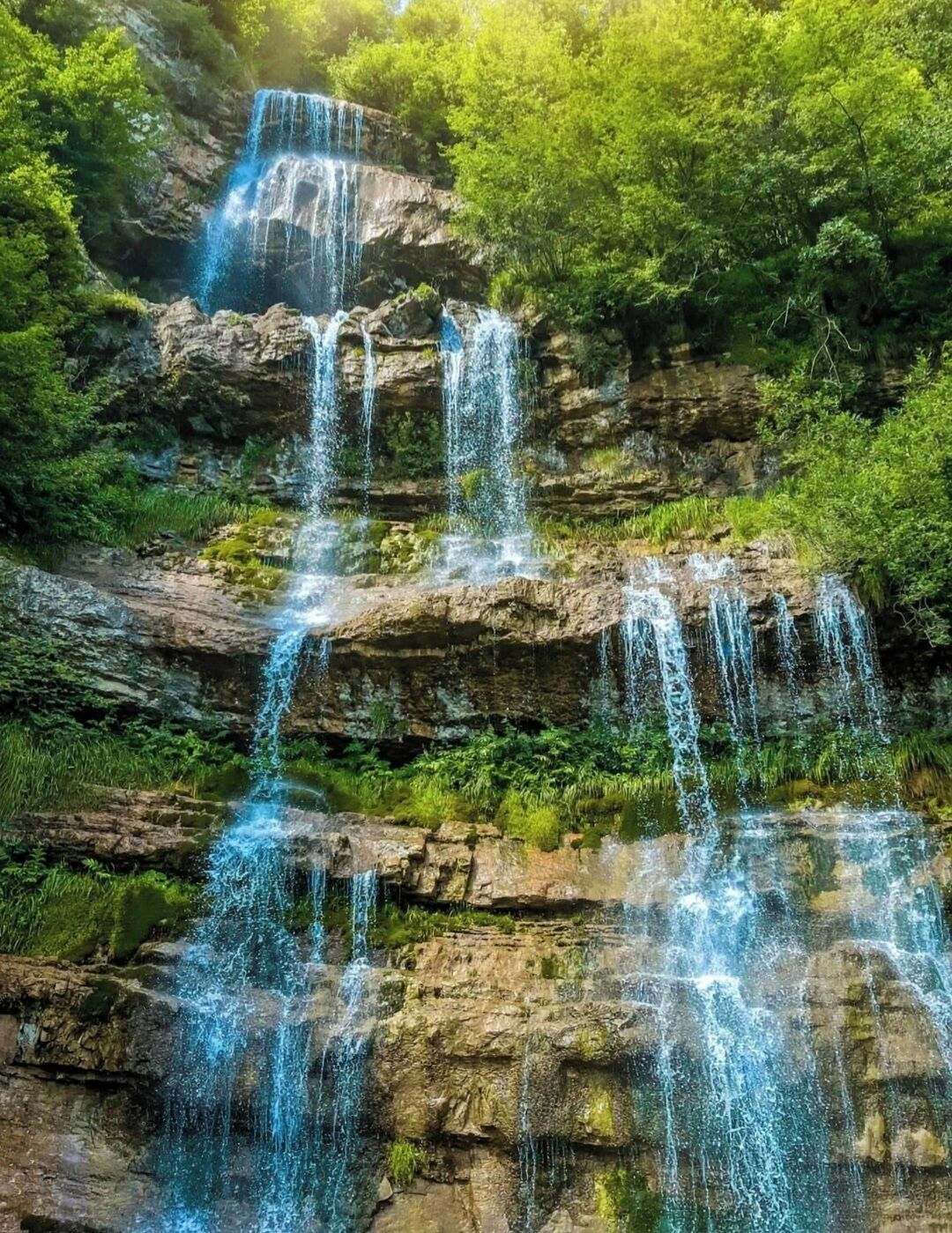 A multi-tiered waterfall cascading over rocky terrain surrounded by lush greenery.