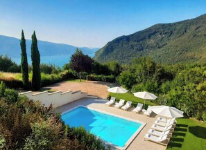 A pool area with lounge chairs and umbrellas, surrounded by greenery and hills.