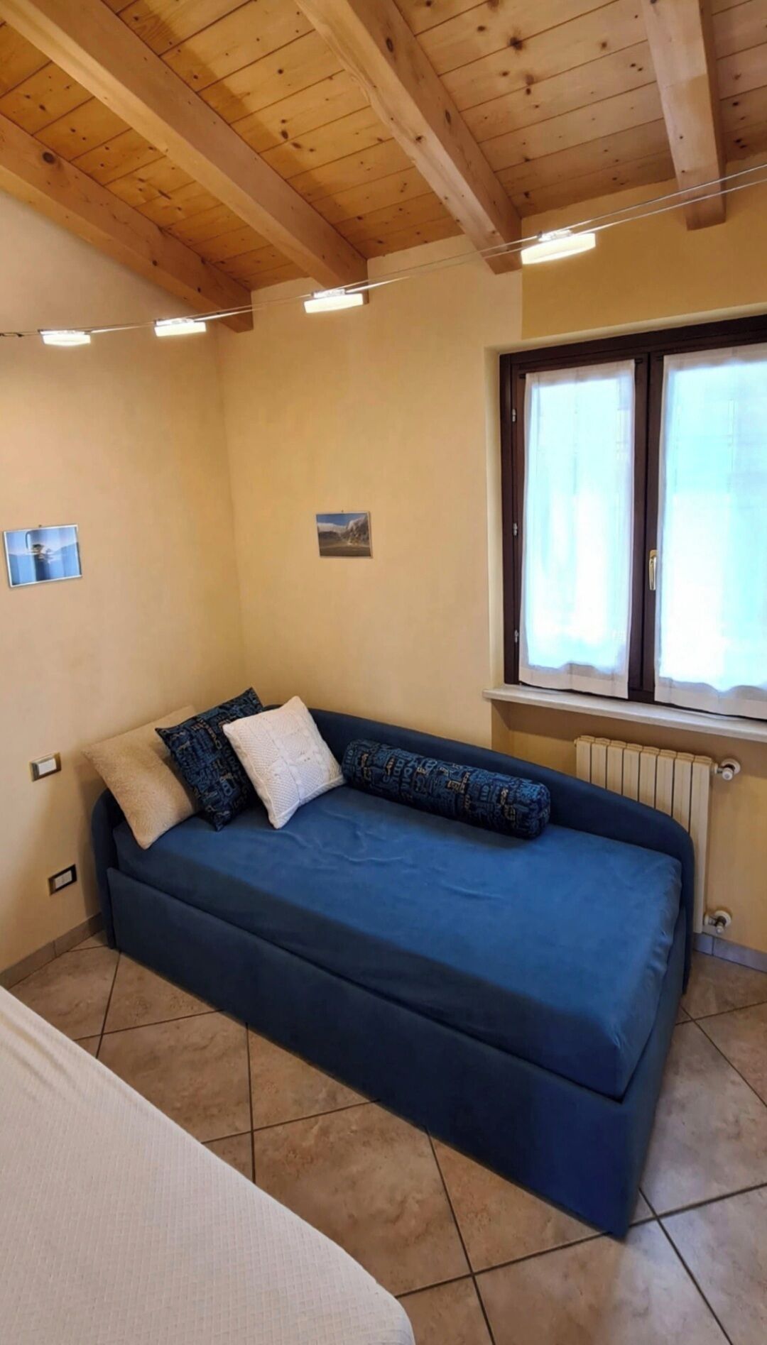 Ein Zimmer mit blauem Sofa, einem Vorhangfenster, einem Heizkörper und einer Holzbalkendecke.