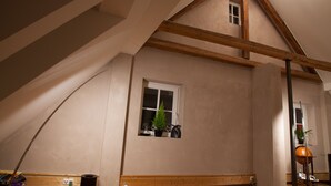 Une pièce avec un plafond en pente, des poutres en bois, une petite fenêtre ornée d’une plante et une lampe.