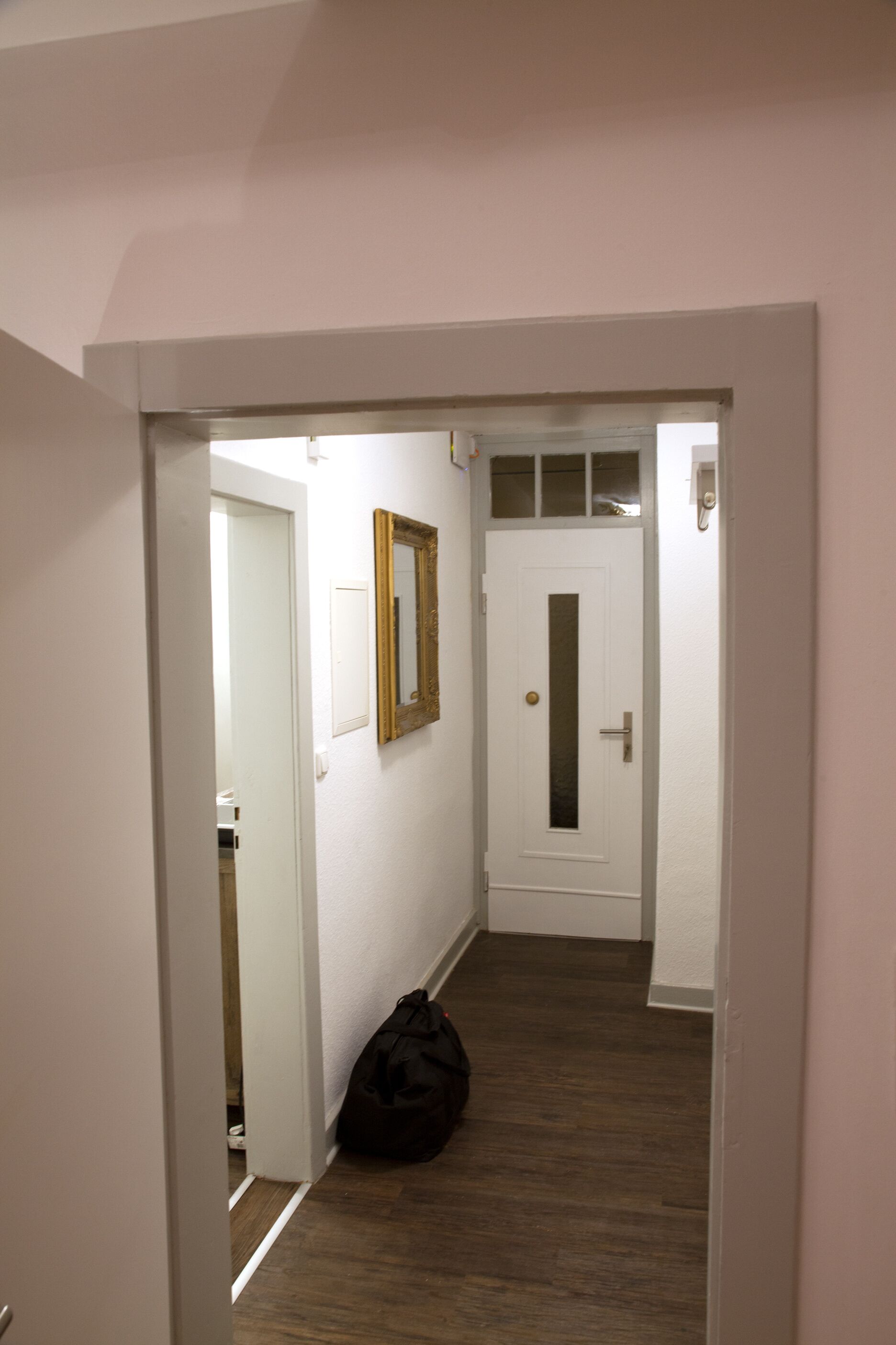 A narrow hallway with a wooden floor, a white door with a small window, and a mirror on the wall.