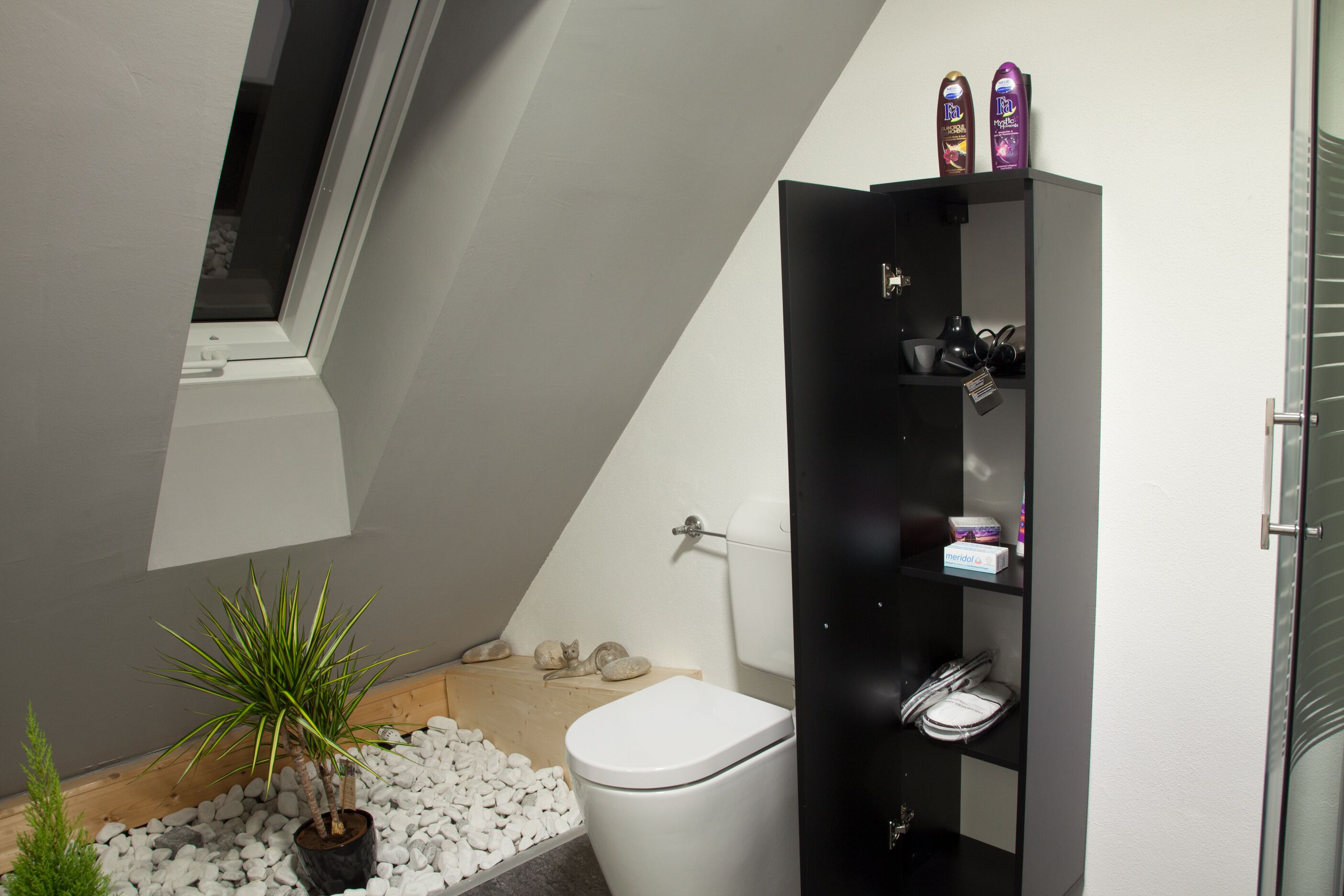 Et moderne badeværelse med et hvidt toilet, et sort skab med toiletartikler og en lille potteplante.
