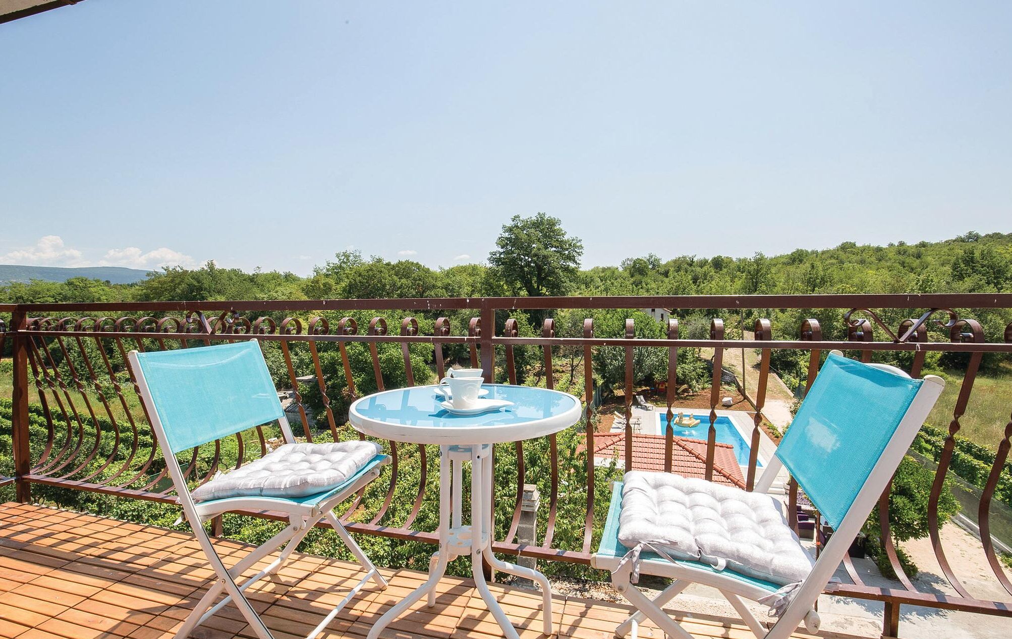 Ein Balkon mit zwei Liegestühlen und einem kleinen Tisch, mit Blick auf ein Schwimmbad und üppiges Grün.