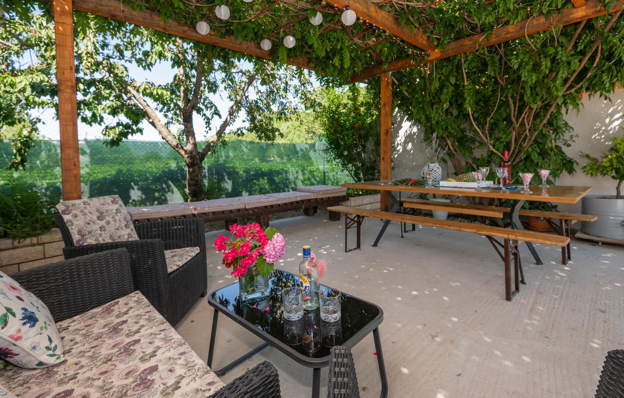 An outdoor patio with wicker furniture, a glass table with drinks, and a pergola with hanging lights.
