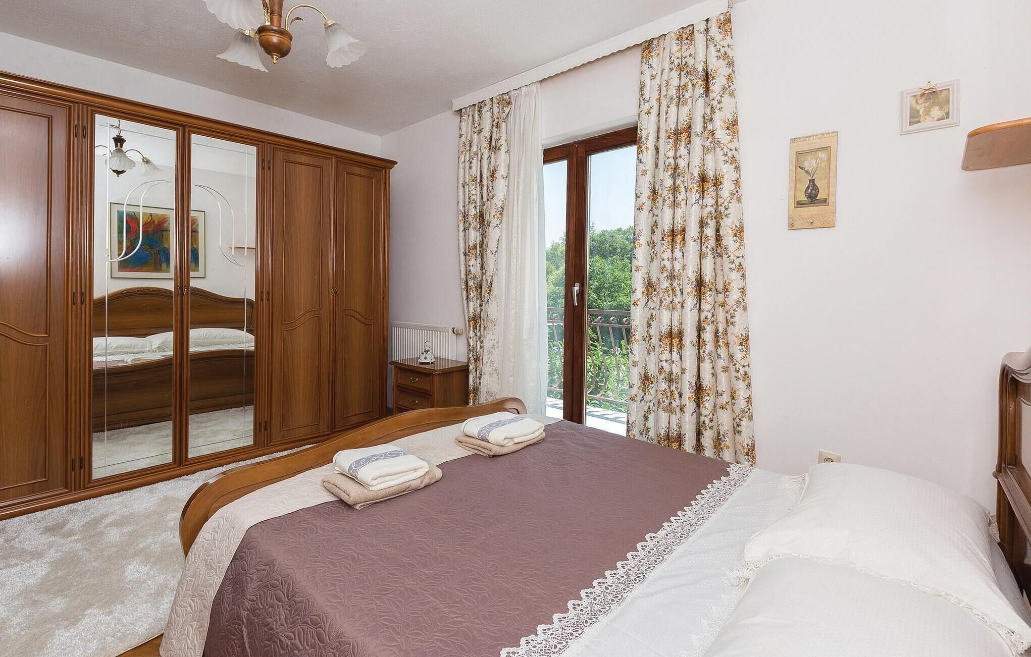 A bedroom with a large wardrobe, a bed with a purple bedspread, and a view of greenery through the window.
