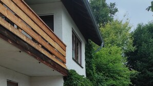 Una casa moderna con balcón, un jardín con rocas y plantas, y mobiliario de exterior.
