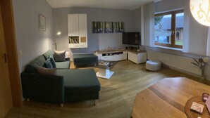 Una sala de estar moderna con un sofá modular, una mesa de centro de vidrio, un televisor y una ventana con vista a una casa.
