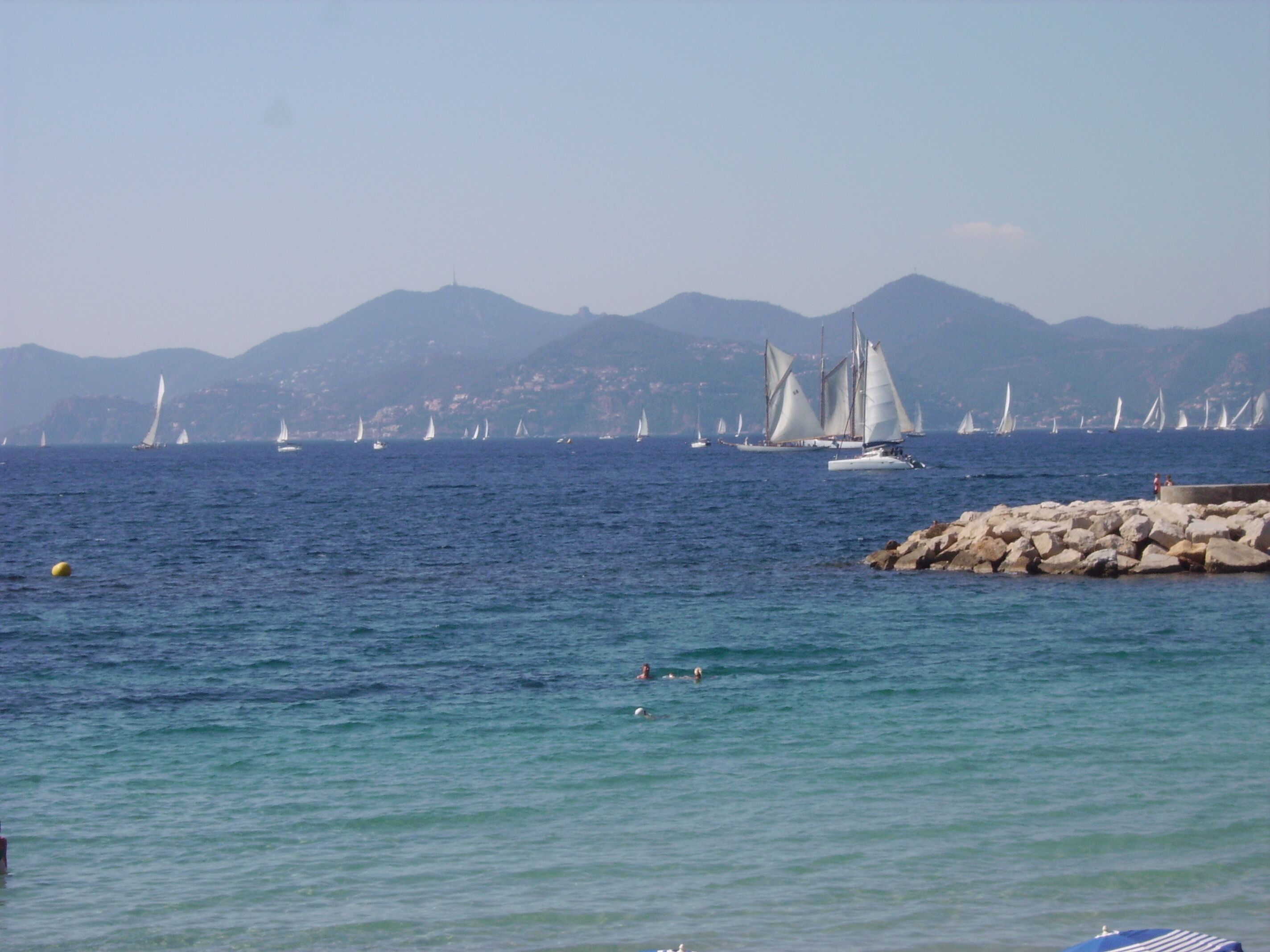 Un paysage côtier avec des voiliers sur l’eau, une côte rocheuse et des montagnes à l’horizon.