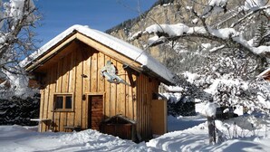 Uma cabana de madeira com telhado inclinado, rodeada por árvores cobertas de neve e uma montanha ao fundo.