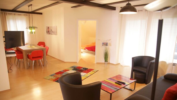 Un salon moderne avec un canapé, une table basse et un coin repas comprenant des chaises et une table. On aperçoit un lit au fond et une plante posée sur le sol.