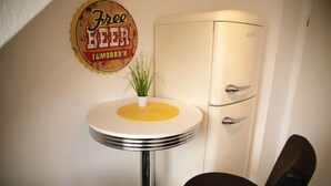 Un coin de pièce avec un réfrigérateur blanc, une petite table blanche surmontée d’un tapis jaune et deux chaises noires.