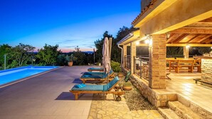 Un área junto a la piscina con sillones de descanso y un porche cubierto con mobiliario exterior.