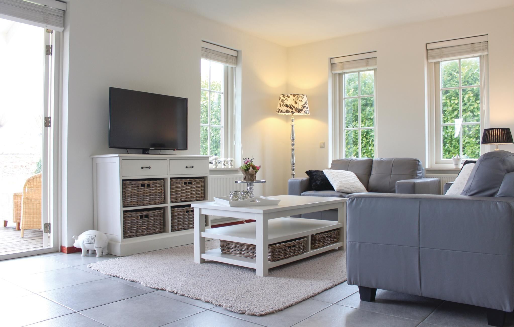 A modern living room with a flat-screen TV, grey sofa, white coffee table, wicker baskets, and a floor lamp.
